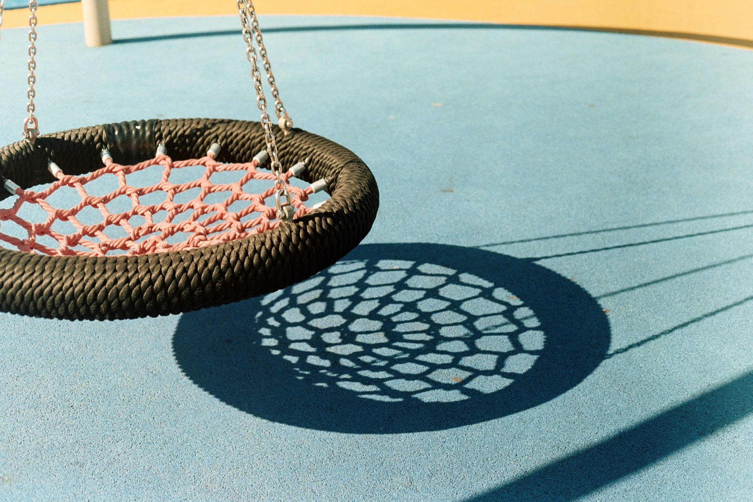 Rope nest swing hanging over smooth blue rubber surfacing, casting a woven shadow