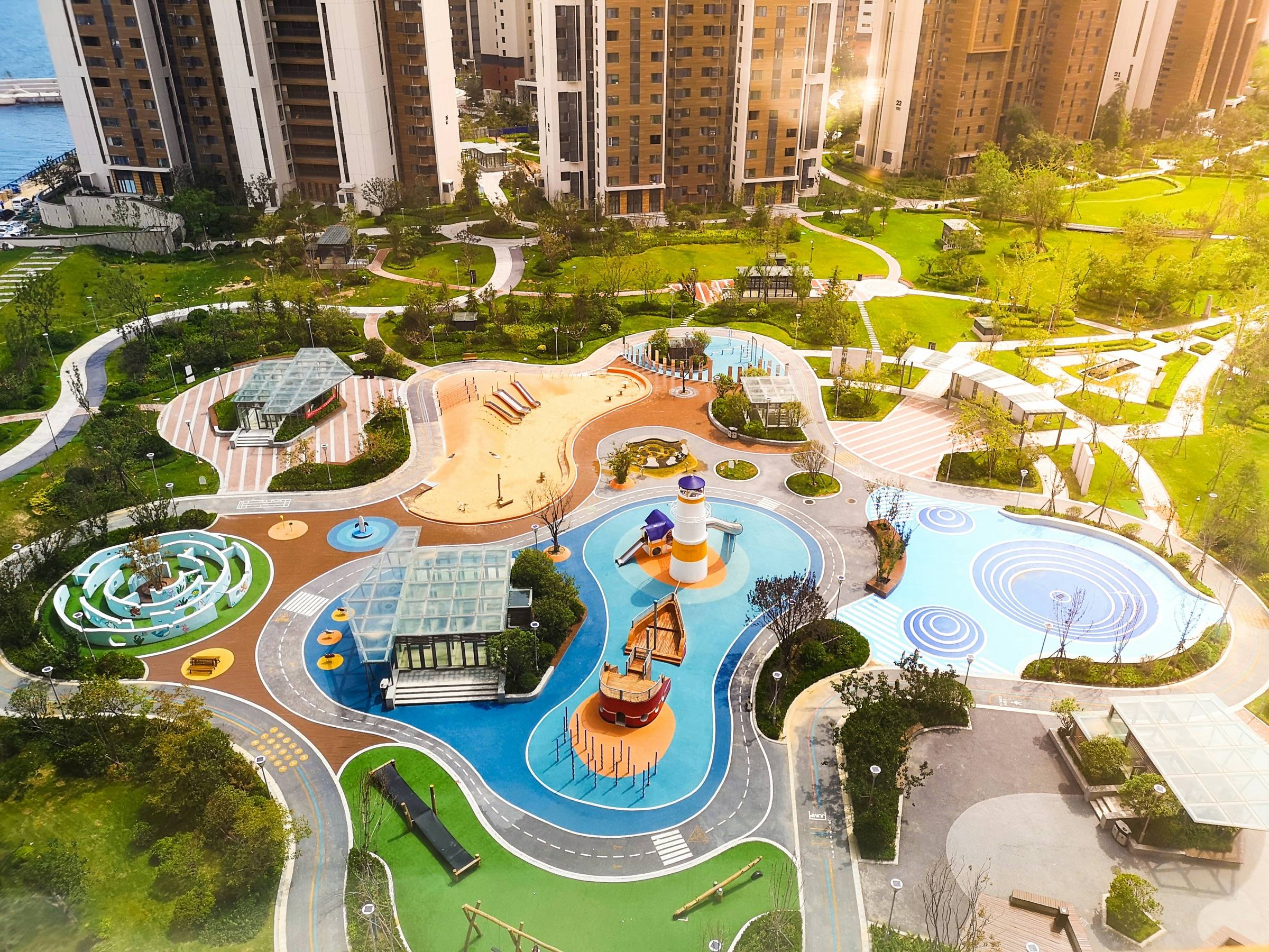 Aerial view of a landscaped community play area with colored surfacing zones, paths, and planting between apartment towers