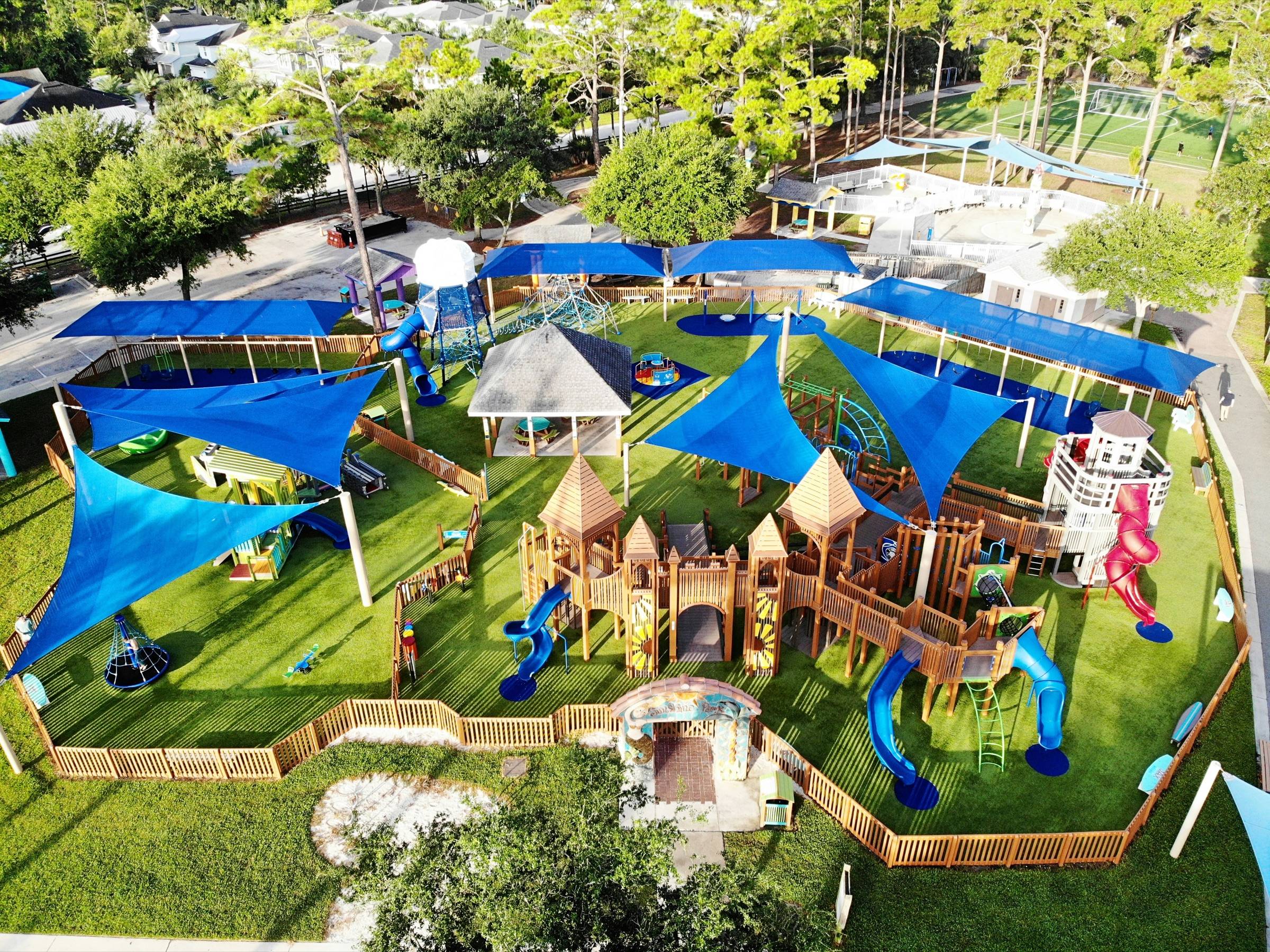Large community playground with blue shade sails over wooden play structures, artificial turf, and a perimeter fence, seen from above